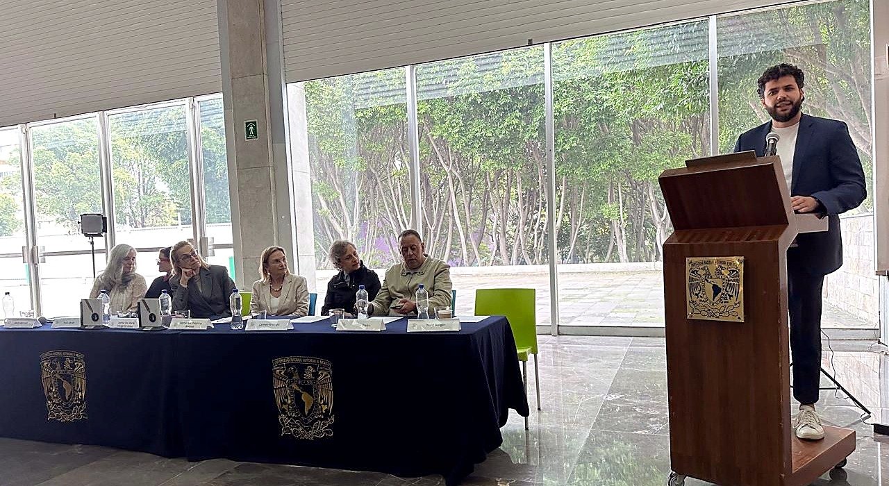 David Boligán habla desde el atril de la UNAM durante la inauguración de la Bienal, con la mesa de invitados a su derecha.